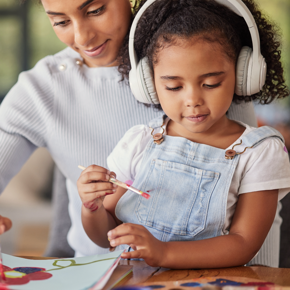 Young girl wearing headphones and painting with mom sitting beside her.